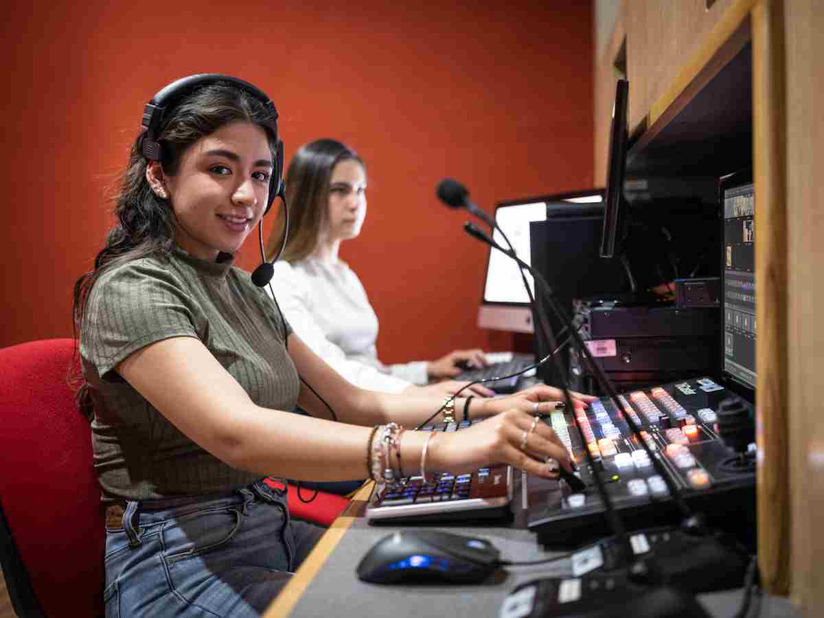 Estudiantes operan una consola de producción con auriculares y micrófonos en una cabina equipada con pantallas y controles de video.
